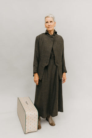 Older white woman, with gray hair wearing the 508 traveling suit, she is standing next to a white suitcase in front of a studio white backdrop