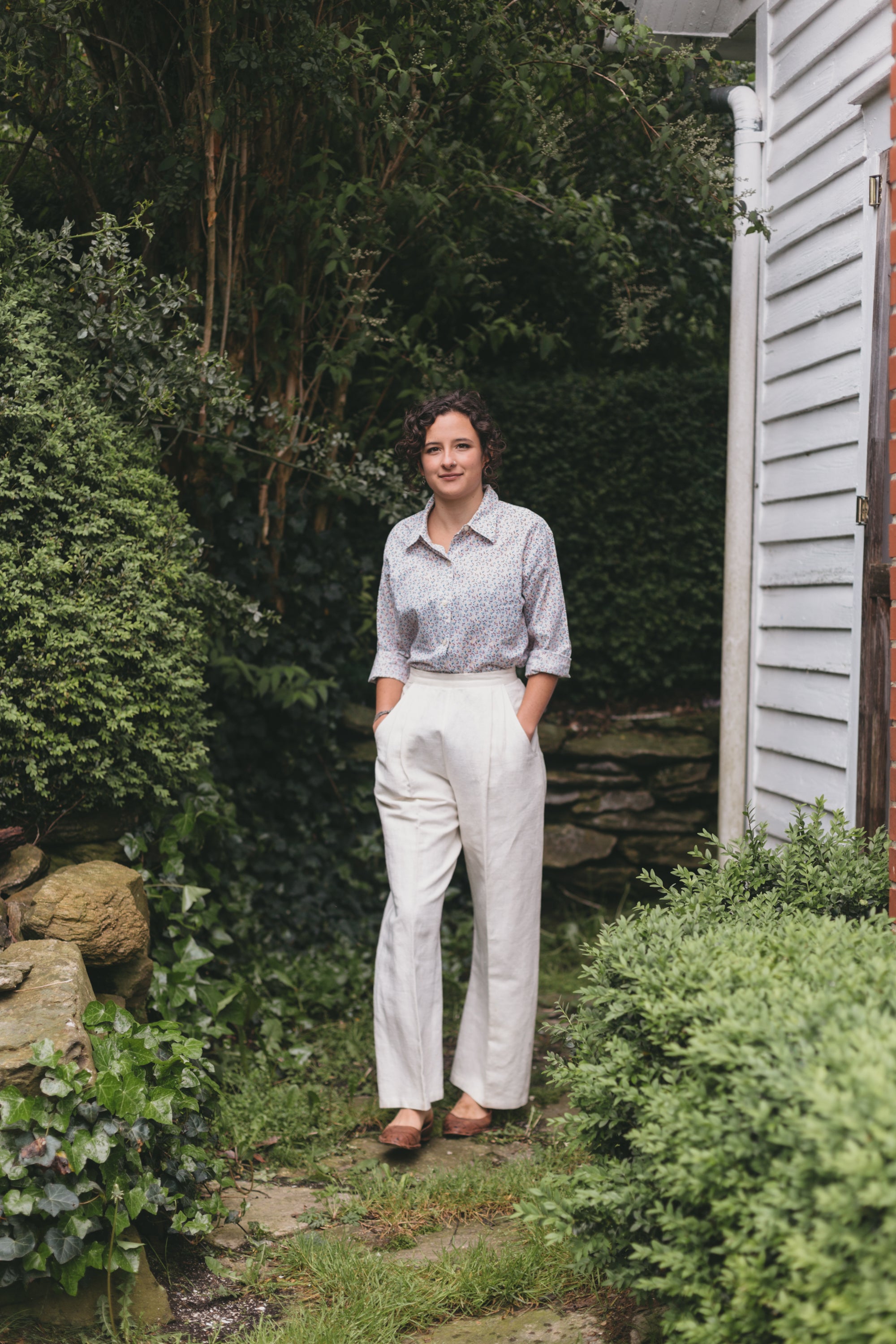 Young brunette white woman walking surrounded by greenery, wearing the Rosie the Riveter slacks and shirt.