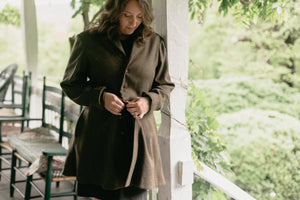 Brunette White woman standing on a porch with greenery in the background buttoning the Countryside Frock Coat.