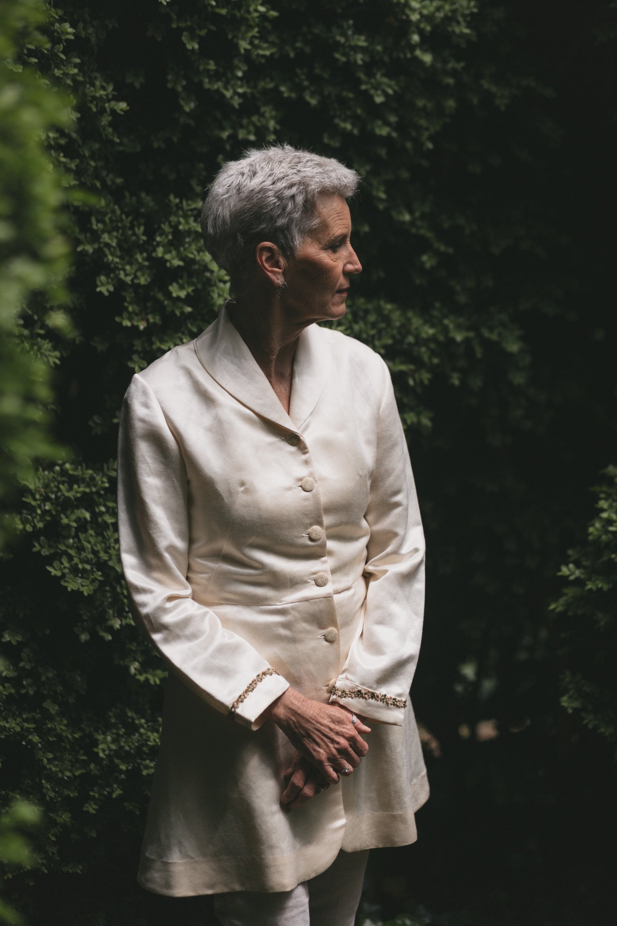 Older white woman with grey hair standing with hands in the front one on top of the other,looking to her left surrounded by greenery wearing the Countryside Frock Coat buttoned in white.