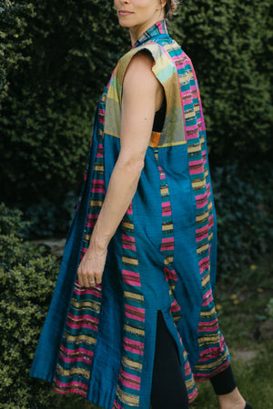 Woman wearing multi colored 118 Tibetan Panel coat.  Standing in front of white background holding sides out in hands.  Woman is looking over her right shoulder showing side slit and shoulder/ arm opening.