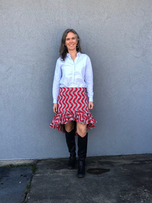 Woman standing in front of a grey wall outside, wearing a buttondown shirt with the Flamenco practice skirt and black boots.  Skirt is red and white ankara fabric and is above the knee in the front and top of calf in the back with a ruffled flounce.