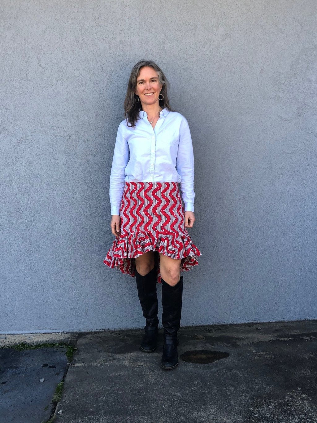 Woman standing in front of a grey wall outside, wearing a buttondown shirt with the Flamenco practice skirt and black boots. Skirt is red and white ankara fabric and is above the knee in the front and top of calf in the back with a ruffled flounce.