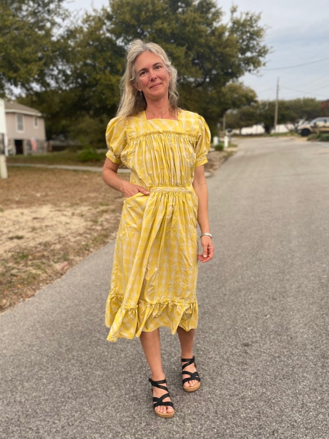 Woman wearing a yellow and white checked calf-length dress with puffed sleeves and pockets.  She is standing in a street and has a hand in her Woman wearing a yellow and white checked calf-length dress with puffed sleeves and pocket.