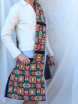 Side view of person holding a bandoleir bag made of black fabric covered in bright yellow, pink, green, and blue embroidery.