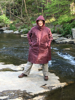 Woman standing on a rock in a creek wearing the parka made of maroon waxed cotton . Her hands are in her pockets.