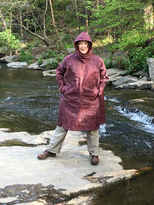Woman standing on a rock in a creek wearing the parka made of maroon waxed cotton .  Her hands are in her pockets.