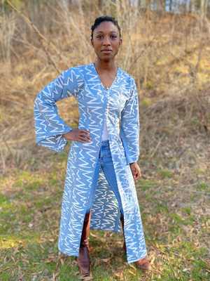 Black woman wearing a long blue/grey batiked entari with brown boots.  She is standing outside.