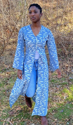 Black woman wearing a long grey blue batiked entari, walking outside.