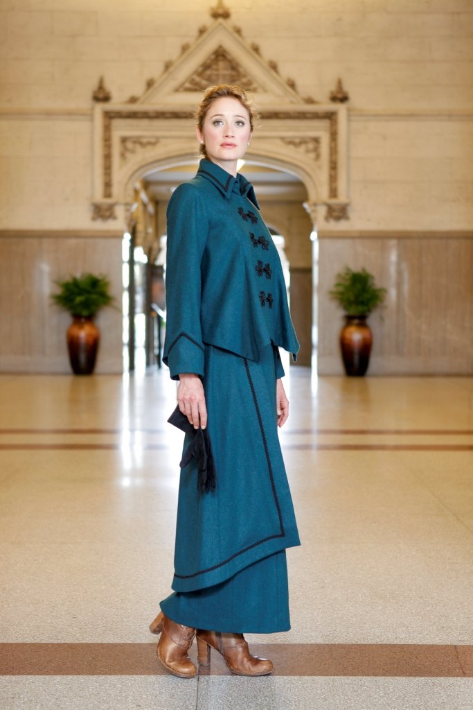 Young blonde woman standing in a lobby wearing the Metropolitan Suit holding black gloves in her right hand and brown heeled boots on her feet.