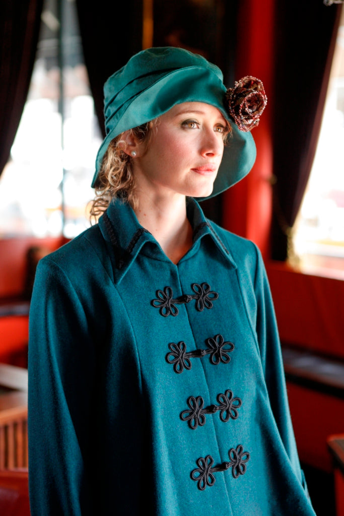 Close up of a young blonde woman in a red lounge area., wearing the Metropolitan suit with front closing frogs, and a hat.