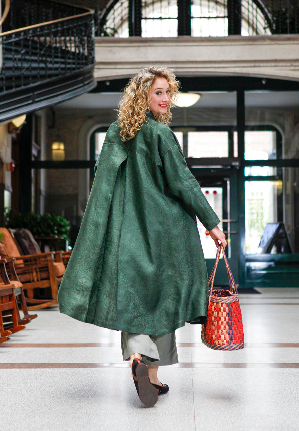 Blonde white woman walking through hallway with her hand in her pocket and holding a woven purse. Wearing the 254 Swing Coat closed with a belt.