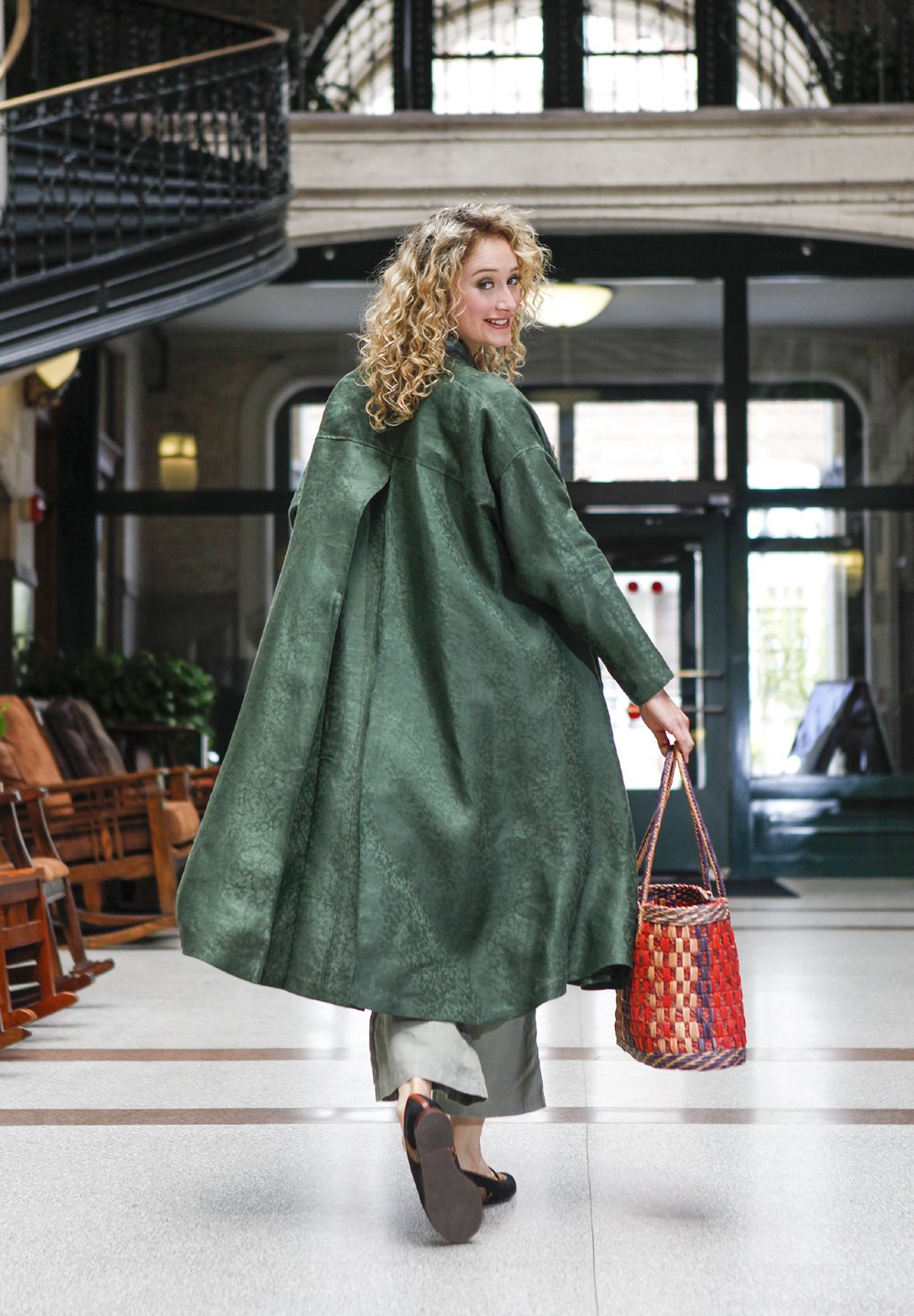 Blonde white woman walking through hallway with her hand in her pocket and holding a woven purse. Wearing the 254 Swing Coat closed with a belt.