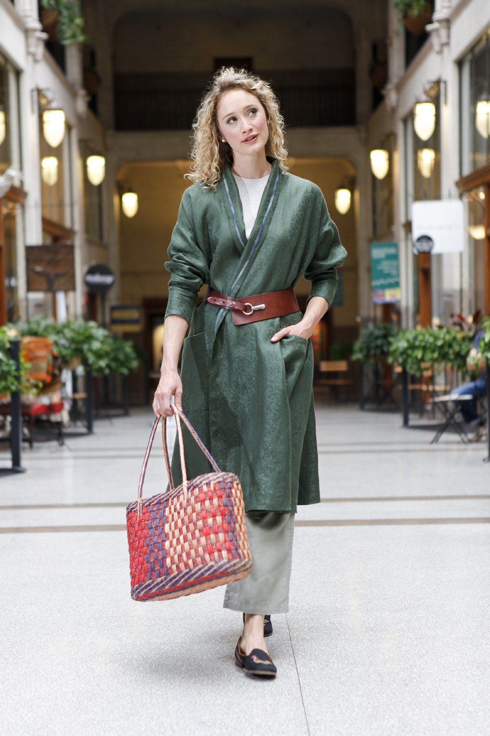 Blonde white woman walking through hallway with her hand in her pocket and holding a woven purse. Wearing the 254 Swing Coat closed with a belt.