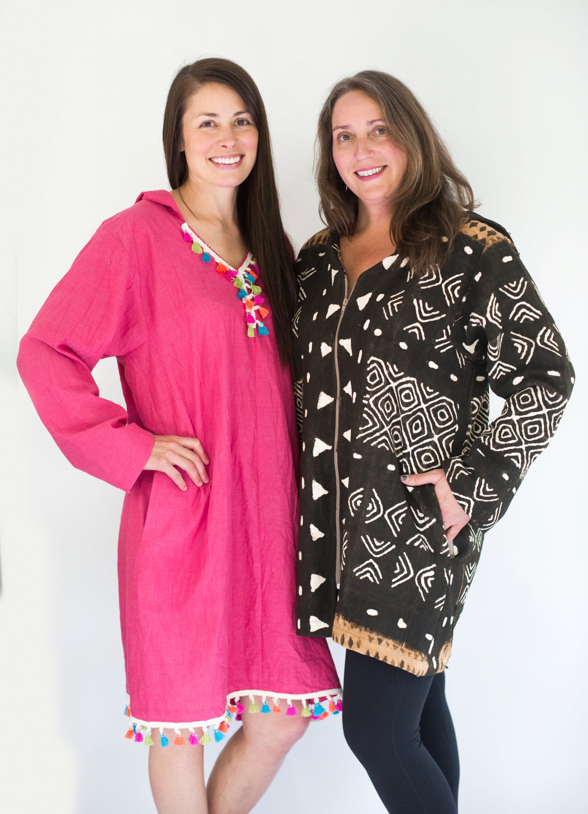 Two Brunette white women standing together smiling with hands on their hips standing in front of a white studio backdrop, wearing the Moroccan Djellaba woman on the left is wearing the pullover while woman on the right is wearing one made with a front zipper. 
